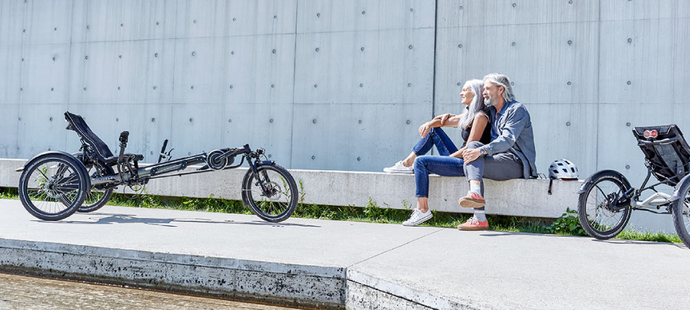 Ein Paar sitzt auf einer niedrigen Mauer daneben stehen die beiden HASE BIKES Dreiräder